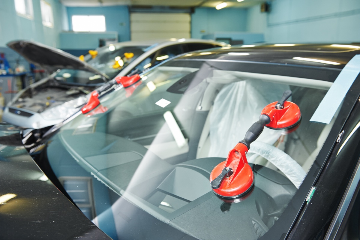 Red suction-cup lifters set on a car's windshield in a repair bay, with a second car behind it, hood up, carrying more lifters.