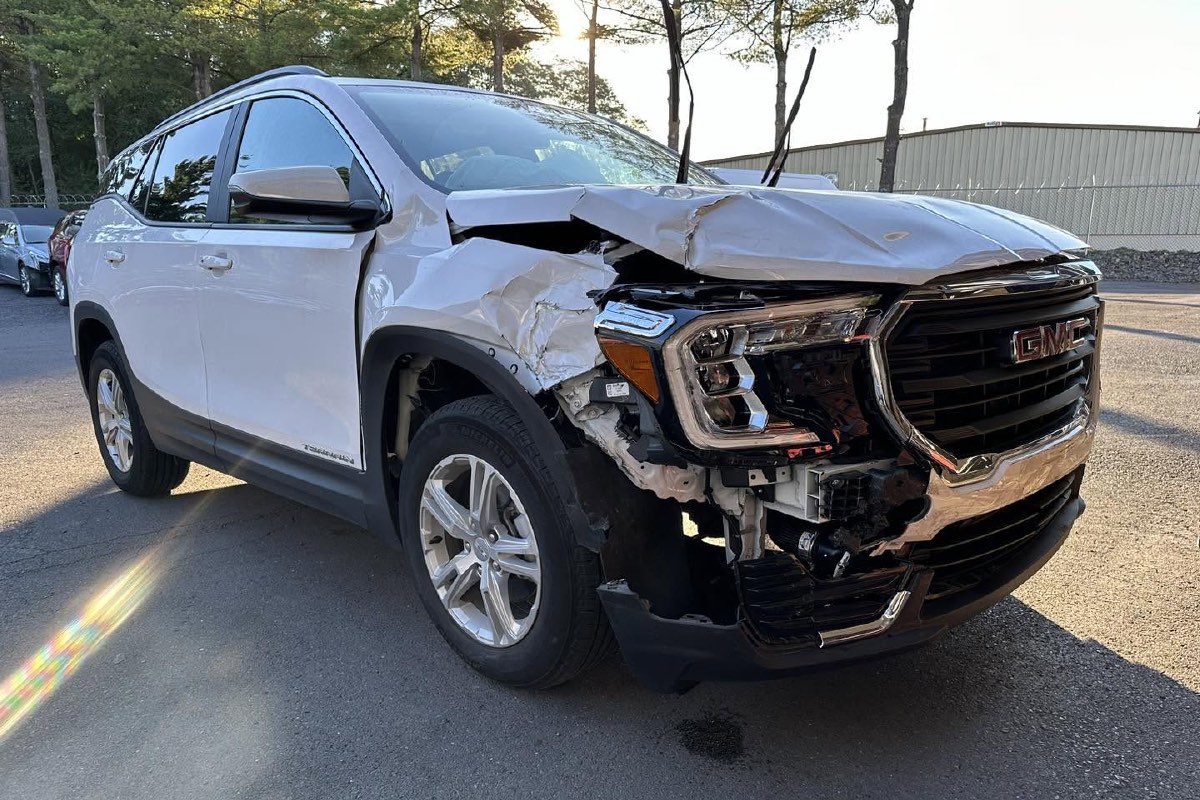 A white GMC SUV with its front end crushed and the headlight torn loose, before repair.