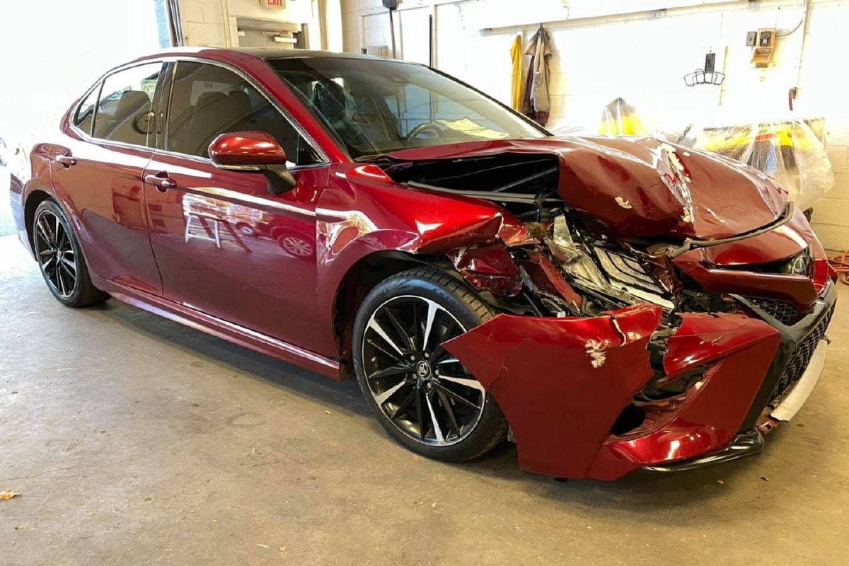 A red sedan with its front end crushed and the bumper torn loose, in the shop before repair.