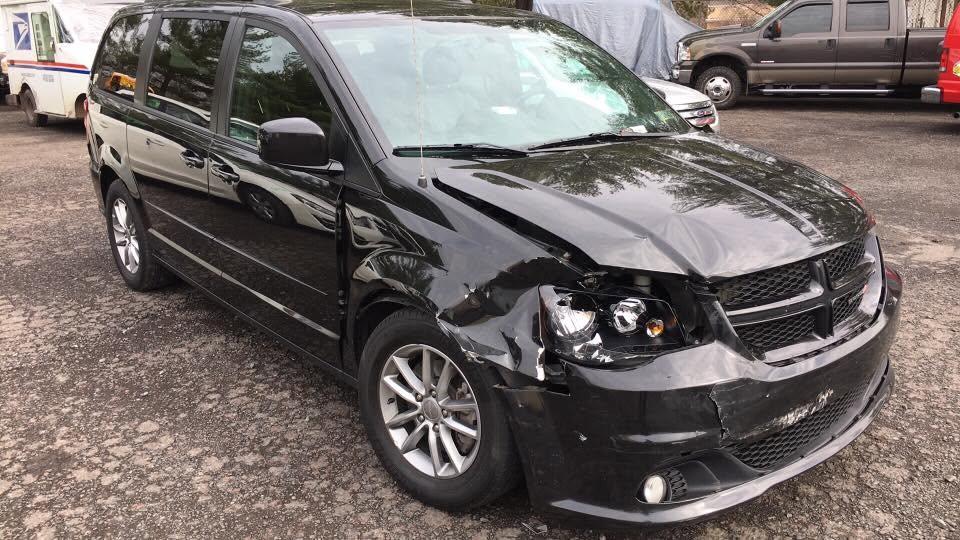 Black Dodge Grand Caravan with a crushed front bumper, a broken headlight and a buckled hood.